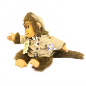 May include: A plush monkey hand puppet with brown fur and a tan face, wearing a tan safari-style jacket with a matching hat. The puppet features a white drawstring, dark buttons, a belt, and a tag that reads PUPPET, plus a circular logo patch.