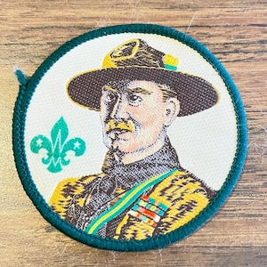 May include: Circular patch featuring a portrait of a man in a hat and uniform, surrounded by a green border. The man has a mustache and is wearing a yellow and green uniform with medals. A green fleur-de-lis is on the left side.