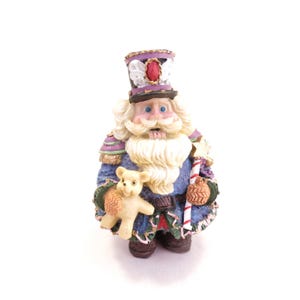 May include: A detailed figurine of a nutcracker soldier, featuring a blue coat with purple and gold accents. The nutcracker holds a teddy bear and a candy cane. The figurine has a white beard and mustache, and a tall hat with a red jewel.