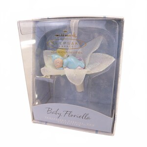 May include: A Hallmark Keepsake Ornament titled Baby Floriella from the Frostlight Fairies, Too collection. The ornament features a small figure in a blue outfit sleeping inside a white, glittery, translucent flower petal structure.