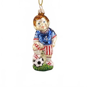 May include: A glass Christmas ornament of a soccer player. The ornament features a boy in a blue jersey with the number 4, red and white striped shorts, and red sneakers. The boy is standing on a soccer ball on a patch of green grass.