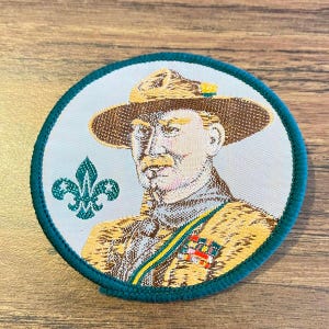 May include: A circular embroidered patch featuring a portrait of a person wearing a wide-brimmed hat and a tan uniform. A green fleur-de-lis emblem is on the left, and a colorful ribbon bar is on the chest. The patch has a dark green border.