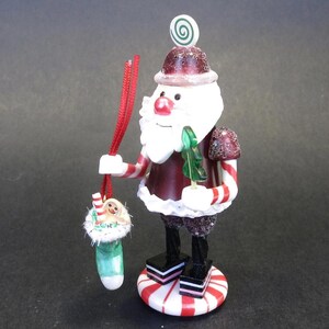 May include: A Christmas ornament of Santa Claus holding a green tree and a stocking with a gingerbread man. The Santa figure has a white beard, red nose, and a red and white striped suit. The ornament is on a red and white striped base.
