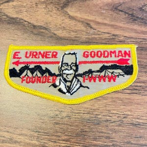May include: A beige embroidered patch with a yellow border featuring a red arrow and the text E. URNER GOODMAN FOUNDER I-WWW. The center displays a black and white illustration of a man with a mustache, positioned above a black mountain silhouette.