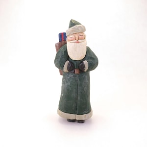 May include: A hand-carved wooden Santa Claus figurine. The figure is dressed in a green coat and hat, with a white beard and a bag of presents on his back. The figurine has a rustic, folk art style.