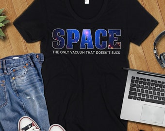 Op de afbeelding: Zwart T-shirt met het woord "SPACE" in een blauw en galaxy-thema lettertype. De tekst "THE ONLY VACUUM THAT DOESN'T SUCK" staat eronder. Het shirt wordt getoond met een spijkerbroek, sneakers, een laptop en een koptelefoon.