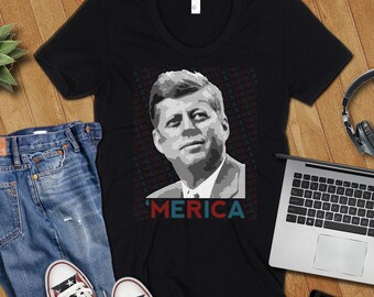Könnte beinhalten: Schwarzes T-Shirt mit einem Graustufenporträt von John F. Kennedy und dem Wort "MERICA" in Rot und Blau. Das Bild zeigt auch eine blaue Jeans, Turnschuhe mit Sternen, einen Laptop, Kopfhörer, ein Smartphone und eine kleine Sukkulentenpflanze.