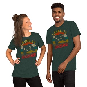 May include: Two t-shirts with a retro-style graphic of a video game controller and the text "Level 21 Unlocked". The shirts are a dark green color.