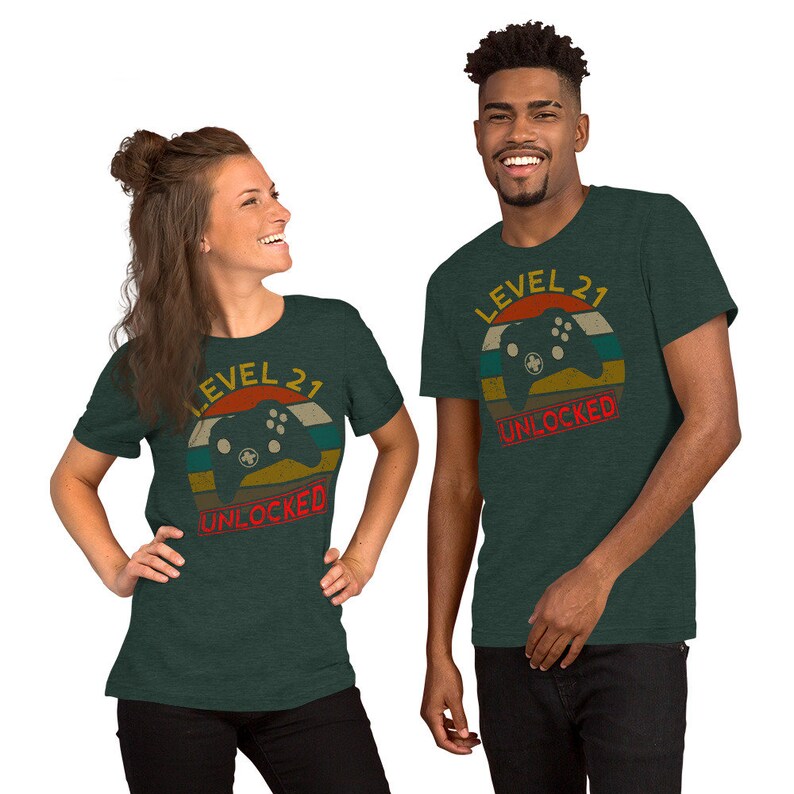 May include: Two t-shirts with a retro-style graphic of a video game controller and the text "Level 21 Unlocked". The shirts are a dark green color.
