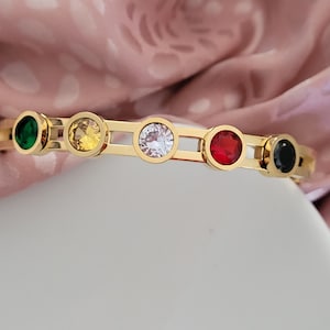 Op de afbeelding: Gouden armband met vijf ronde edelstenen: groen, geel, wit, rood en zwart.
