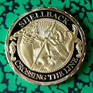May include: A gold-colored coin with a black background. The coin features a depiction of a mythical sea creature with a trident and the words "Shellback Crossing the Line".