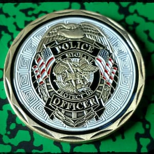 May include: Gold-colored police officer challenge coin with a black and white design. The coin features an eagle with wings spread, a shield with the words "Police", "St. Michael", "Protect Us", and "Officer", and two American flags.