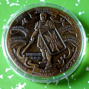 Puede incluir: Una moneda de bronce con la imagen de un soldado con armadura, sosteniendo una espada y un escudo. La moneda tiene el texto "Ponte toda la armadura de Dios" y "Efesios 6:11-13" alrededor del borde.