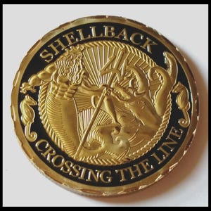 May include: A gold-colored coin with black accents features a detailed relief of a mythical sea god with a trident battling a sea creature. The coin's edge is inscribed with "SHELLBACK CROSSING THE LINE" in a circular pattern, with seahorses on the sides.