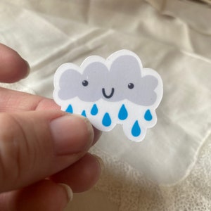 May include: A small, white sticker in the shape of a smiling cloud with gray shading and blue raindrops. The cloud has a happy face with black eyes and a curved black mouth. The sticker is held between fingers.