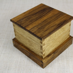 Puede incluir: Una caja de madera con una tapa de color marrón oscuro y una base de color marrón claro. La caja está hecha de madera con un patrón de veta visible. La tapa está ligeramente elevada sobre la base.