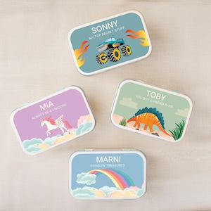 May include: Four rectangular metal tins with rounded corners, each featuring a unique graphic design and name. Designs include a blue monster truck, a purple unicorn, a green dinosaur, and a blue sky with a rainbow and clouds.