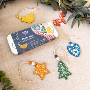 May include: A Cotton Twist Folk Art Decoration Kit in a rectangular tin box with five wooden ornaments including a yellow bird, red horse, blue heart, orange star, and green tree, each with a string and wooden beads for hanging.