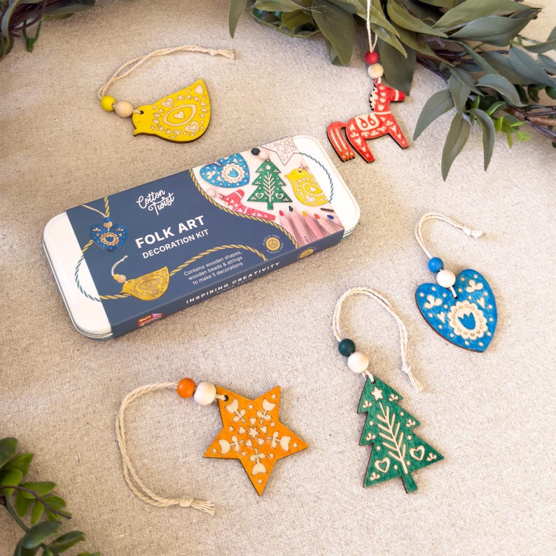 May include: A Cotton Twist Folk Art Decoration Kit in a rectangular tin box with five wooden ornaments including a yellow bird, red horse, blue heart, orange star, and green tree, each with a string and wooden beads for hanging.