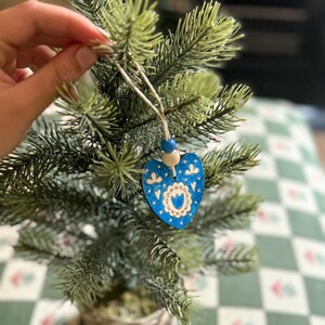 May include: A heart-shaped wooden ornament featuring a blue painted surface with intricate white floral and geometric patterns. The ornament is suspended by a string adorned with one blue and one natural wooden bead, hanging from a green pine branch.