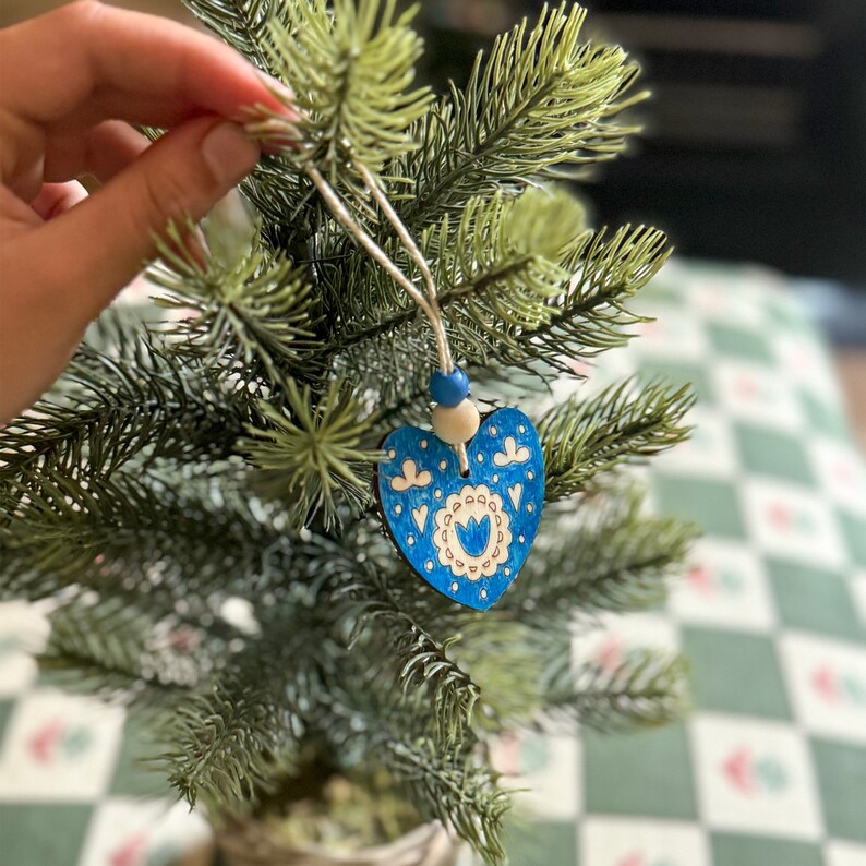 May include: A heart-shaped wooden ornament featuring a blue painted surface with intricate white floral and geometric patterns. The ornament is suspended by a string adorned with one blue and one natural wooden bead, hanging from a green pine branch.