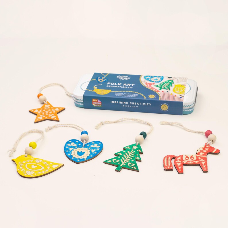 May include: A Folk Art decoration kit in a rectangular tin box with blue, white, and yellow branding. Included are five wooden ornaments: a yellow star, a yellow bird, a blue heart, a green tree, and a red horse, each with a string and beads.