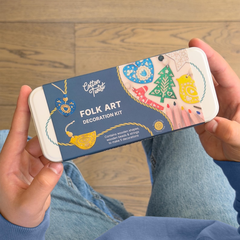 May include: A rectangular white metal tin featuring a blue paper sleeve labeled Cotton Twist Folk Art Decoration Kit. The sleeve displays illustrations of five colorful wooden ornaments, including a blue heart, green tree, and yellow bird.