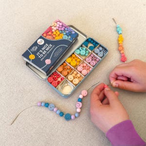 Könnte beinhalten: Ein Armband-Bastelset mit einer Vielzahl bunter Perlen, darunter Smiley-Gesichter, in einer geteilten Blechdose. Das Set enthält den Text "IT'S NICE TO BE NICE BRACELET BEADING KIT". Ein teilweise fertiges Armband ist abgebildet.