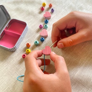 May include: A craft kit with colorful beads and heart-shaped wooden pieces with red and white stripes. A needle is being used to thread a blue string through a heart bead. A small, striped tin is open in the background.