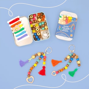 May include: A keyring making kit in a metal tin featuring wooden beads in various shapes and colors, colorful thread, needles, and metal clips. The kit includes text reading You & Me Keyring Making Kit. Two finished keyrings with beads and tassels are shown.