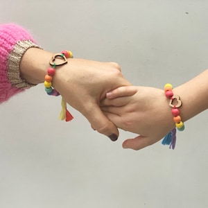 May include: Two matching bracelets featuring multicolored wooden beads, a central wooden heart-shaped charm, and small hanging tassels. The beads are arranged in a rainbow color sequence, and the bracelets are worn on two separate wrists.