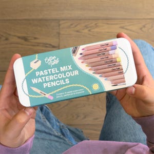 May include: A rectangular metal pencil case with a light blue background and the words "PASTEL MIX WATERCOLOUR PENCILS". The case contains 10 pastel watercolour pencils, a paintbrush, and a sharpener. The case is held by a person.