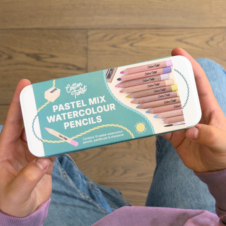 May include: A rectangular metal pencil case with a light blue background and the words "PASTEL MIX WATERCOLOUR PENCILS". The case contains 10 pastel watercolour pencils, a paintbrush, and a sharpener. The case is held by a person.