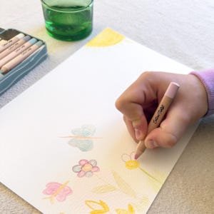 May include: A hand drawing a flower with a pink crayon on white paper. The paper features a sun, flowers, and a butterfly drawing. A green glass and a pencil case with crayons are in the background. The crayon is labelled "Cotton Twist".