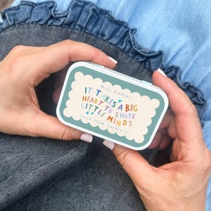 May include: A rectangular metal tin with a teal scalloped border and a cream-coloured centre. The text reads: Miss Gadsby, IT TAKES A BIG HEART TO SHAPE LITTLE MINDS, Love from Saskia x. The tin features small blue heart accents.