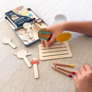 May include: A DIY solar system craft kit featuring wooden planet cutouts, a rectangular wooden base with the text THE SOLAR SYSTEM, coloured pencils, and a metal storage tin. The kit includes various planet shapes to colour and assemble.