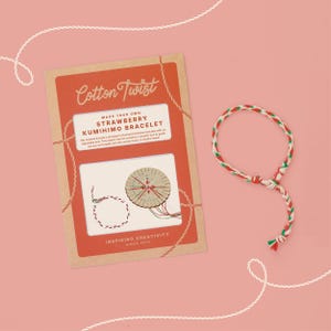 Op de afbeelding: Een Cotton Twist Make Your Own Strawberry Kumihimo Bracelet knutselpakket in een rechthoekige kartonnen verpakking met rode en bruine accenten, inclusief een rond houten vlechtgereedschap en een afgewerkte rood, wit en groen gevlochten armband.