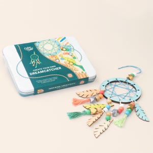 May include: A craft kit in a rectangular white tin labelled Cotton Twist Create Your Own Dreamcatcher, featuring a teal band with white text. Beside it lies a completed dreamcatcher with a teal wooden ring, blue string web, and hanging wooden beads, tassels, and wooden feathers in shades of teal, orange, yellow, and grey.