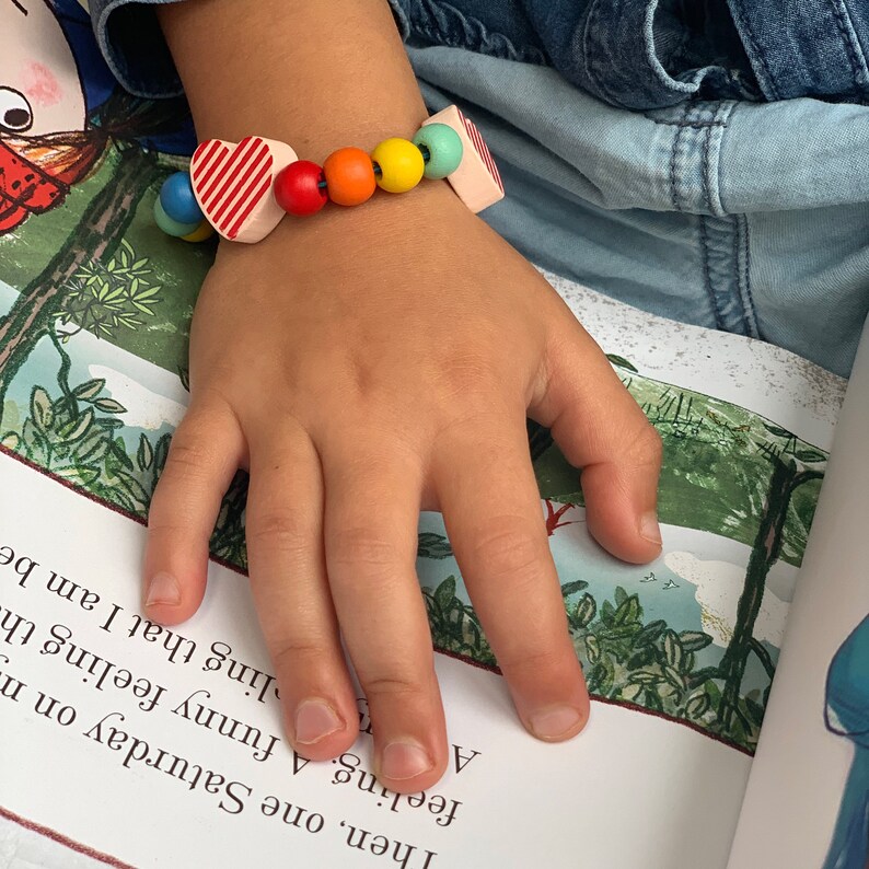 May include: A colorful wooden bead bracelet with a heart and square-shaped beads. The bracelet features red, orange, yellow, and green beads. The heart-shaped bead has red and white stripes. The bracelet is on a child's hand.