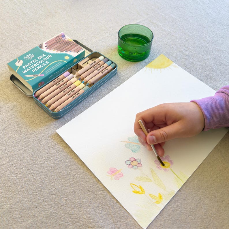 May include: A teal tin of pastel mix watercolour pencils is open, next to a drawing in progress. The pencils are arranged in rows. A hand holds a paintbrush, adding colour to a floral design. A green glass of water is also present.