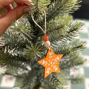 May include: A star-shaped wooden ornament featuring an orange finish with white floral patterns. The ornament hangs from a twine loop accented with one orange bead and one white bead, suspended from the branch of an artificial pine tree.