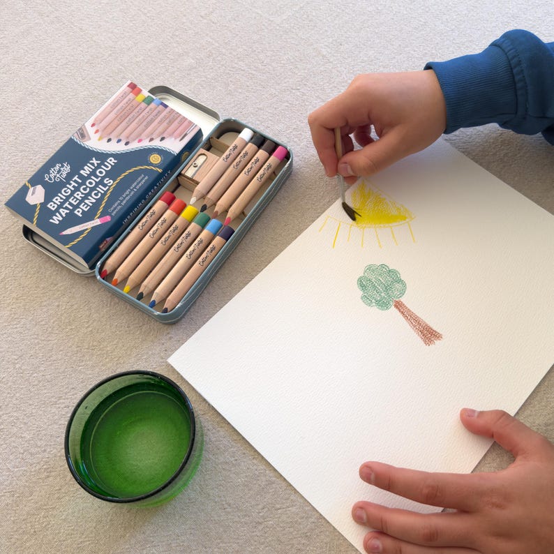 May include: A set of watercolor pencils in a blue tin, with the text "Bright Mix Watercolour Pencils". The pencils are arranged with a pencil sharpener. A child is drawing a sun and tree on white paper with the pencils. A green glass is in the foreground.