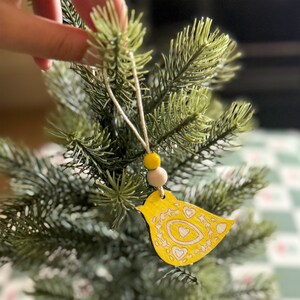 May include: A yellow wooden bird ornament featuring intricate engraved heart and floral patterns. The bird hangs from a thin string adorned with one bright yellow bead and one natural light-colored wooden bead, suspended from a green pine branch.