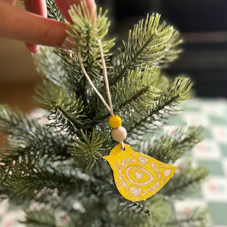 May include: A yellow wooden bird ornament featuring intricate engraved heart and floral patterns. The bird hangs from a thin string adorned with one bright yellow bead and one natural light-colored wooden bead, suspended from a green pine branch.