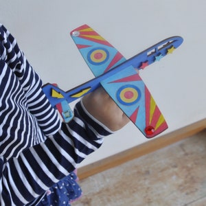 May include: A colorful wooden toy airplane with red, yellow, blue, and white stripes. The plane has a red and yellow circle on each wing and a red jewel on the tail.