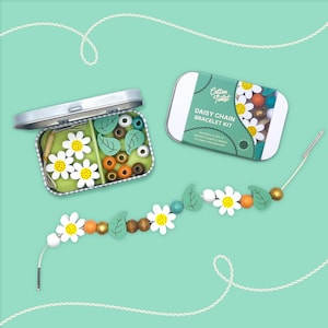 Könnte beinhalten: Ein Daisy Chain Armband-Set mit einem fertigen Armband, Perlen und Werkzeugen. Das Set enthält weiße, margeritenförmige Perlen mit gelben Zentren, grüne Blattperlen und verschiedene andere farbige Perlen. Das Set ist in einer Metalldose verpackt.