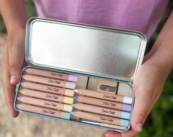Puede incluir: Estuche rectangular azul claro abierto, con lápices de colores pastel y un sacapuntas. Los lápices, etiquetados como "Cotton Twist", son de varios tonos pastel.