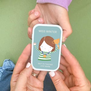 May include: A rectangular metal tin with a light blue background featuring a cartoon illustration of a person with brown hair wearing a green and white striped shirt, holding a gold trophy, and wearing a gold medal with the text MISS WINTER BEST PE TEACHER LOVE MARNI.