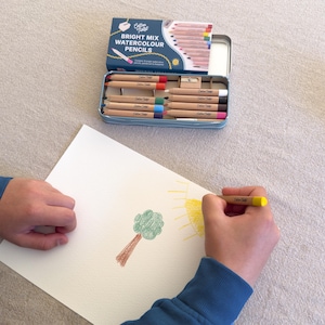 May include: A blue tin of Cotton Tales Bright Mix Watercolour Pencils is open, revealing a set of colored pencils. A person is drawing a tree and sun with the pencils on white paper. The tin has a white lid with text.
