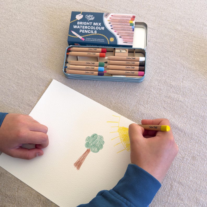 May include: A blue tin of Cotton Tales Bright Mix Watercolour Pencils is open, revealing a set of colored pencils. A person is drawing a tree and sun with the pencils on white paper. The tin has a white lid with text.